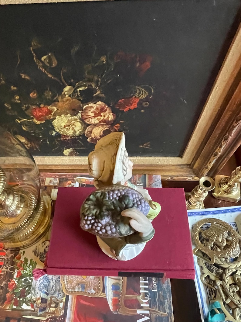 K&ouml;nnte beinhalten: Keramikfigur einer Person, die eine Traube h&auml;lt. Die Figur hat einen beigen Hut und einen wei&szlig;en Sockel. Die Trauben sind lila und gr&uuml;n. Die Figur steht auf einem roten Buch.