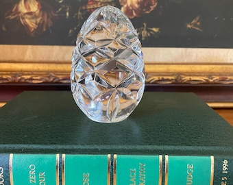 May include: A clear, faceted glass egg-shaped decorative object. The egg is sitting on a green book with gold lettering. The book titles include "The Zero Hour", "Rose", "A Place for Kathy", and "The Judge".