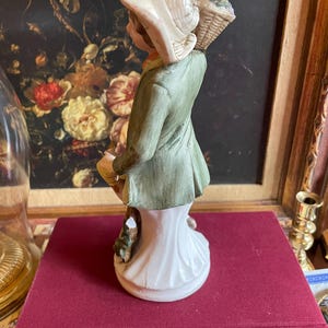 K&ouml;nnte beinhalten: Keramikfigur einer Person mit Hut und Korb. Die Figur tr&auml;gt einen gr&uuml;nen Mantel und eine wei&szlig;e Basis. Hut und Korb sind beige. Die Figur steht auf einem roten Buch.