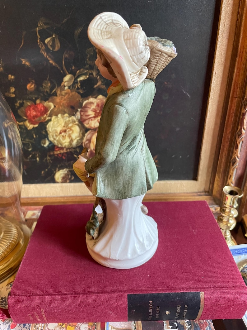 K&ouml;nnte beinhalten: Keramikfigur einer Person mit Hut und Korb. Die Figur tr&auml;gt einen gr&uuml;nen Mantel und eine wei&szlig;e Basis. Hut und Korb sind beige. Die Figur steht auf einem roten Buch.