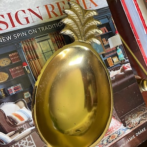 May include: A polished gold-toned metal dish shaped like a pineapple. The bowl features a smooth, concave interior and a textured, leafy top section. The metallic surface reflects light, highlighting the curved edges and the detailed pineapple leaves.