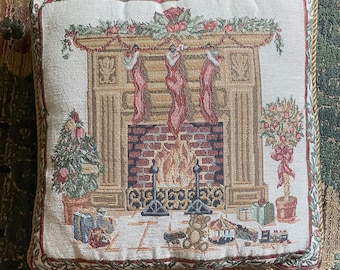 May include: A square throw pillow featuring a woven tapestry design of a brick fireplace with a roaring fire, three hanging red stockings, a decorated Christmas tree, a potted topiary, and various wrapped gifts on the floor, finished with gold trim.