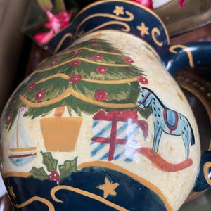 May include: A ceramic Christmas pitcher with a cream-colored background and a navy blue rim. The pitcher is decorated with a Christmas tree, presents, a rocking horse, and a sailboat. The pitcher has a handle and a spout.