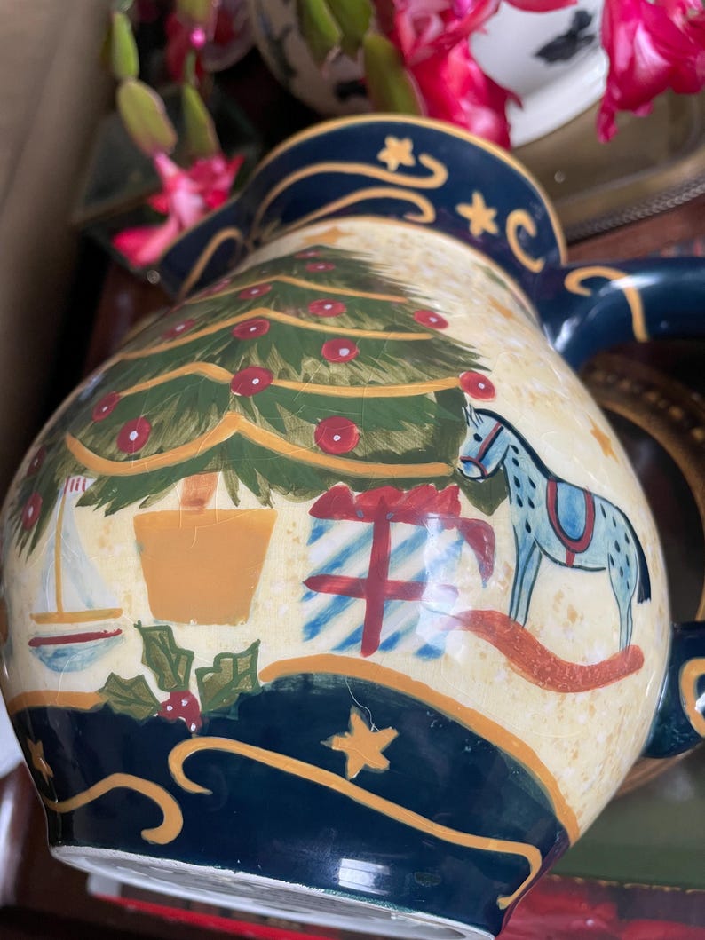 May include: A ceramic Christmas pitcher with a cream-colored background and a navy blue rim. The pitcher is decorated with a Christmas tree, presents, a rocking horse, and a sailboat. The pitcher has a handle and a spout.