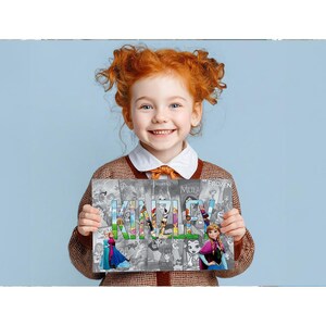 A young girl with curly red hair is smiling and holding up a sign with the name "KINLEY" written on it.