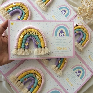 May include: A pink, yellow, green, and purple rainbow made of yarn with gold butterfly accents and a gold name tag that says "Rosa". The rainbow is on a white card with a pink background and a text message in Italian that says "Ogni bimba parla con se un arcobaleno, parte dal cuore ed esce verso il cielo con un sorriso. Rosa. Il mio 1 Compleanno. 13 Novembre 2022."