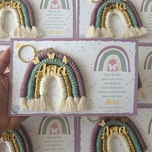 Op de afbeelding: Een set van vijf regenboogkleurige sleutelhangers met de naam "Aria" in gouden letters. Elke sleutelhanger is bevestigd aan een kaart met een boodschap in het Italiaans die luidt: "Ogni bimba porta con se un arcobaleno, parte dal cuore ed esce verso il cielo con un sorriso. Aria Il mio Santo Battesimo 8. Ottobre 2022".
