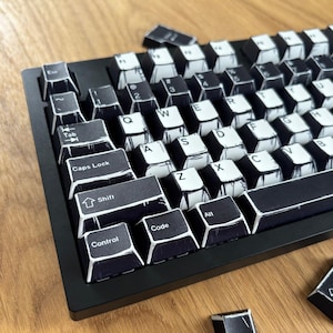 May include: A black and white mechanical keyboard with keycaps featuring a hand-drawn aesthetic. The keyboard sits on a wooden surface, with several detached keycaps scattered around. The keycaps have letters, numbers, and symbols.