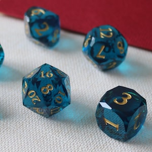 May include: A set of seven polyhedral dice in various shapes, with a translucent teal color and gold numbering. The dice are arranged on a white surface, with a red and beige background.