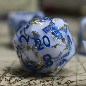 May include: A set of blue and white dice with gold accents. The dice are made of a translucent material and have a swirling pattern. The numbers on the dice are in blue and white.