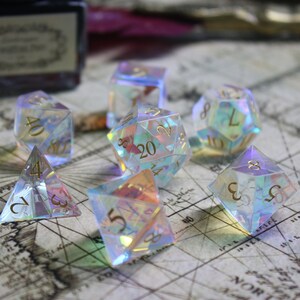 May include: A set of iridescent dice with gold numbers on a vintage map background. The dice are made of clear plastic and have a rainbow effect. The dice are a d4, d6, d8, d10, d12, and d20.