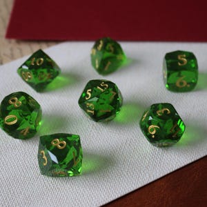 Puede incluir: Juego de siete dados poliédricos verdes con numeración dorada. Los dados se muestran sobre una superficie blanca texturizada, con un sobre rojo y otros artículos de papelería en el fondo. Dados para juegos de mesa.