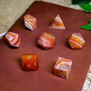 May include: Seven agate dice in various shapes and sizes, with gold numbers, on a brown leather journal. The dice are a mix of red, orange, and white.