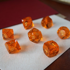 Puede incluir: Conjunto de siete dados poliédricos naranjas translúcidos con numeración dorada. Los dados están dispuestos sobre una superficie blanca, con un fondo rojo y una superficie similar al cuero en el fondo. Para juegos de rol.
