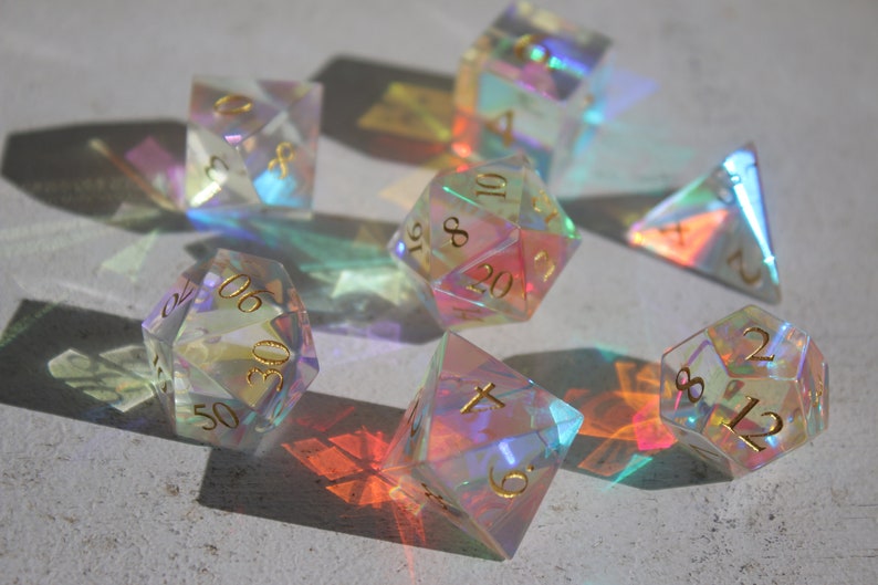 May include: A set of iridescent dice with gold numbers. The dice are made of clear plastic and have a rainbow effect. The dice are in various shapes, including a pyramid, a cube, and a 20-sided die.