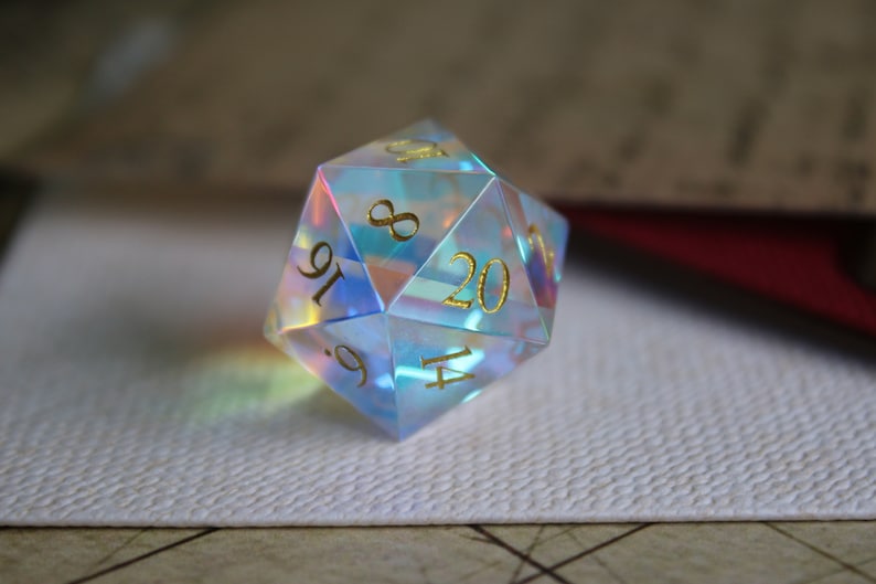 May include: A clear iridescent 20-sided die with gold numbers. The die is sitting on a white surface.
