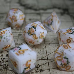 May include: Set of seven polyhedral dice with a floral design. The dice are white with gold numbering and feature pink and green floral accents. The dice are designed for role-playing games.