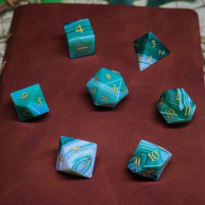 Könnte beinhalten: Satz von sieben grünen und weißen Achatwürfeln mit goldenen Zahlen auf einer braunen Lederoberfläche. Die Würfel sind ein W4, W6, W8, W10, W%, W12 und W20.