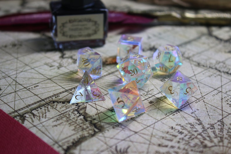 May include: A set of iridescent dice with gold numbers on a vintage map background. The dice are made of clear plastic and have a rainbow effect. The dice are a d4, d6, d8, d10, d12, and d20.