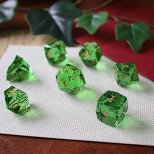 Puede incluir: Se muestran siete dados verdes facetados con números dorados. Los dados están dispuestos sobre una superficie blanca, con un fondo rojo y hojas verdes. Los dados son de varias formas y tamaños.