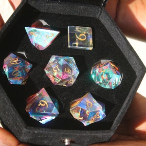 May include: A set of seven iridescent dice in a black velvet box. The dice are made of clear glass with gold numbers and have a rainbow effect. The dice are in a hexagonal box with a black velvet lining.