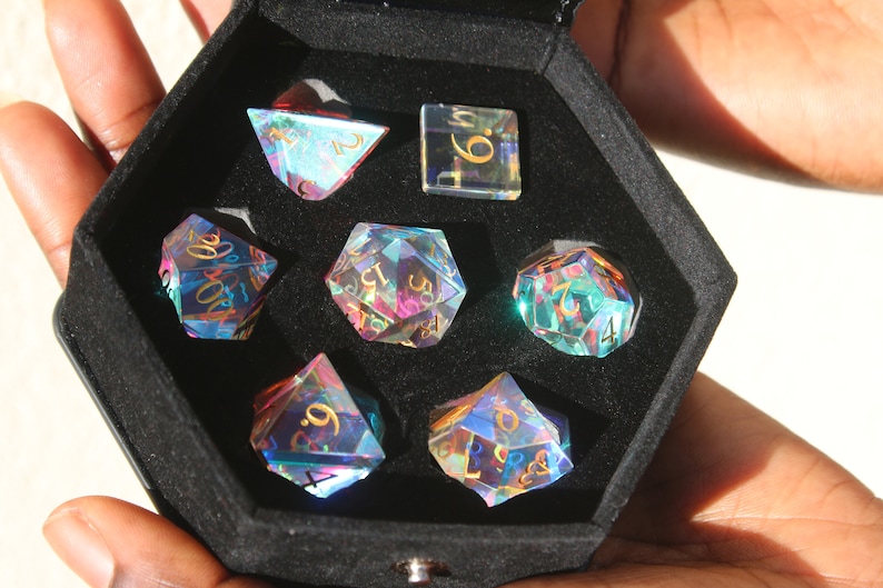 May include: A set of seven iridescent dice in a black velvet box. The dice are made of clear glass with gold numbers and have a rainbow effect. The dice are in a hexagonal box with a black velvet lining.