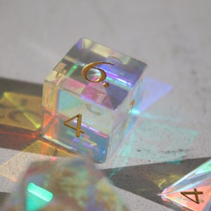 May include: A set of iridescent dice with gold numbers. The dice are made of clear plastic and have a rainbow effect. The dice are showing the numbers 4 and 6.