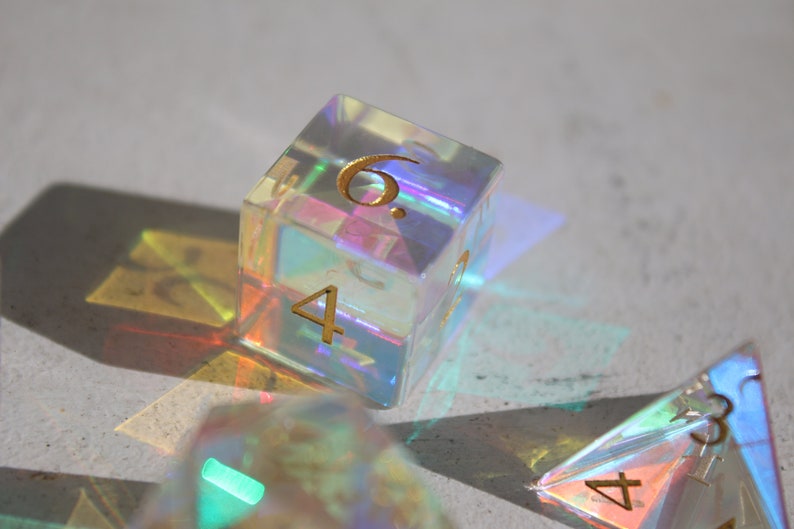May include: A set of iridescent dice with gold numbers. The dice are made of clear plastic and have a rainbow effect. The dice are showing the numbers 4 and 6.