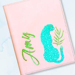 Op de afbeelding: Een roze notitieboek met een turquoise luipaardontwerp en groene accenten. Het woord "Amy" is in groene letters geschreven. De tekst "LEOPARD JOKER TURQUOISE, GREEN, WITH GOLD ACCENTS" is ook zichtbaar.