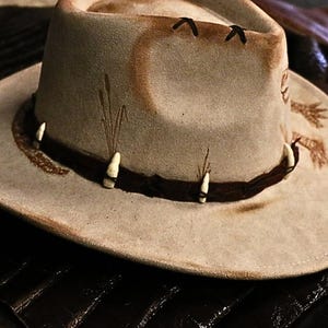 Pu&ograve; includere: Cappello da cowboy beige chiaro con fascia in pelle marrone scuro e dettagli decorativi. Il cappello presenta un'ampia tesa e un aspetto leggermente usurato, con dettagli dipinti e piccoli abbellimenti bianchi.