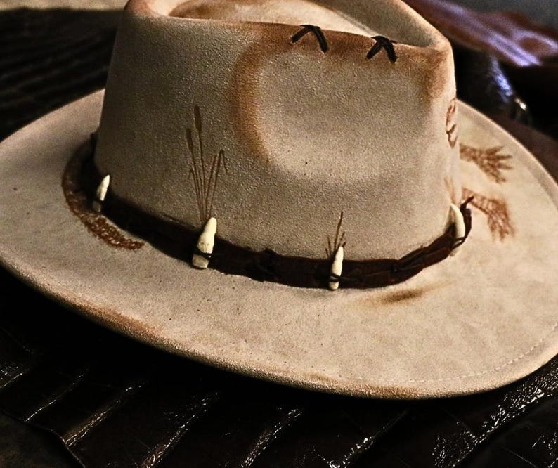 Pu&ograve; includere: Cappello da cowboy beige chiaro con fascia in pelle marrone scuro e dettagli decorativi. Il cappello presenta un'ampia tesa e un aspetto leggermente usurato, con dettagli dipinti e piccoli abbellimenti bianchi.