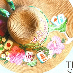 May include: A wide-brimmed straw hat with a tropical theme. The hat features a margarita glass, hibiscus flowers, and Mahalo text. Decorative elements include lime slices and Mahjong tiles. The hat has a green and white patterned ribbon.