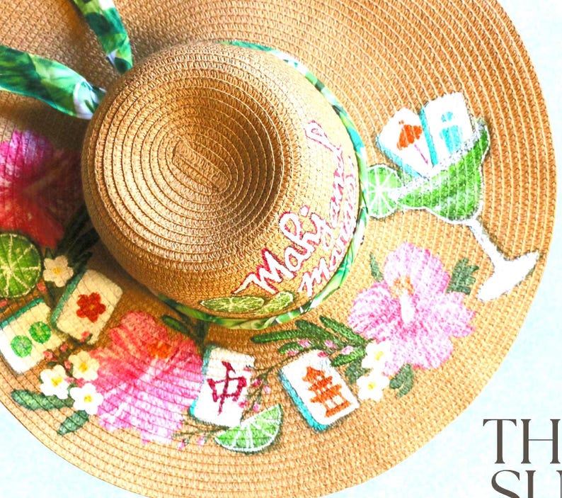 May include: A wide-brimmed straw hat with a tropical theme. The hat features a margarita glass, hibiscus flowers, and Mahalo text. Decorative elements include lime slices and Mahjong tiles. The hat has a green and white patterned ribbon.