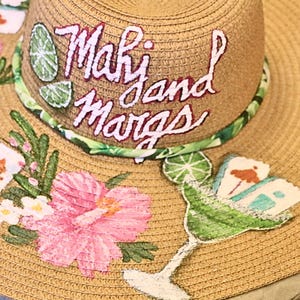 May include: A wide-brimmed straw hat with the words "Mahi and Margs" embroidered in white. The hat is decorated with pink hibiscus flowers, green lime slices, and a margarita glass design. The hat has a green and white patterned band.