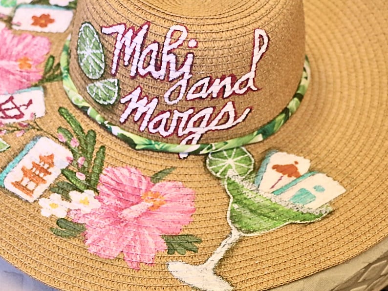 May include: A wide-brimmed straw hat with the words "Mahi and Margs" embroidered in white. The hat is decorated with pink hibiscus flowers, green lime slices, and a margarita glass design. The hat has a green and white patterned band.