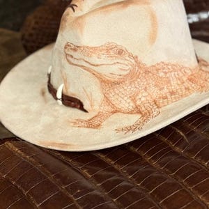 Pu&ograve; includere: Un cappello da cowboy beige con una dettagliata illustrazione marrone di un alligatore. Il cappello ha un'ampia tesa e una fascia in pelle marrone con elementi decorativi. Il cappello &egrave; posto su una superficie marrone strutturata.