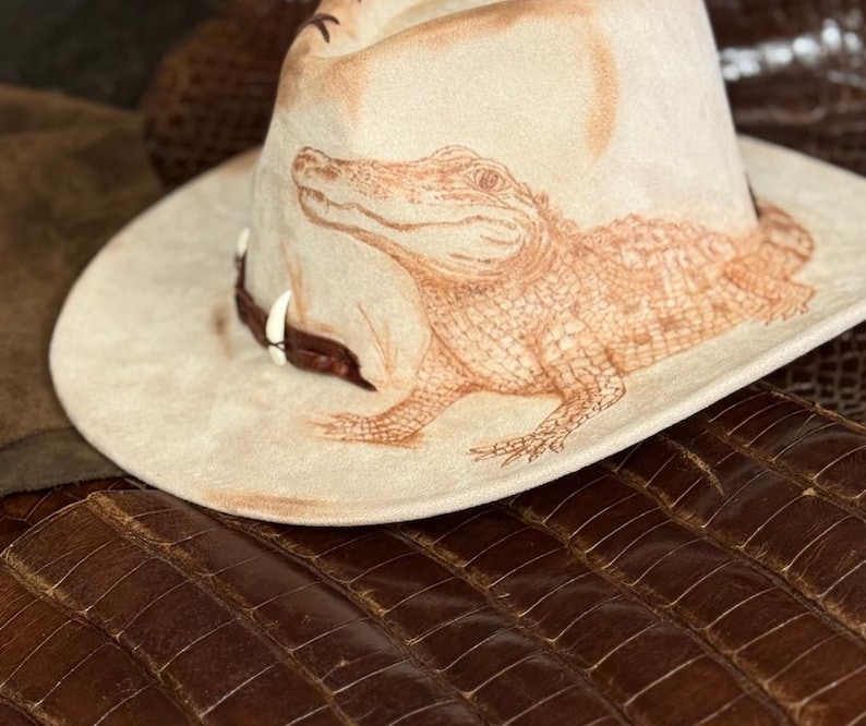 Pu&ograve; includere: Un cappello da cowboy beige con una dettagliata illustrazione marrone di un alligatore. Il cappello ha un'ampia tesa e una fascia in pelle marrone con elementi decorativi. Il cappello &egrave; posto su una superficie marrone strutturata.