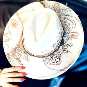 May include: A cream-colored fedora hat with a sun and moon design etched in brown. The hat features a decorative band and a classic brim. The sun and moon artwork adds a celestial touch to this stylish accessory.