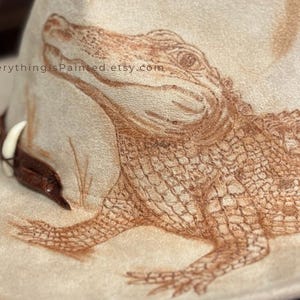 Pu&ograve; includere: Un cappello da cowboy beige con una dettagliata illustrazione marrone di un alligatore. Il disegno dell'alligatore &egrave; finemente disegnato sulla corona del cappello. Il cappello ha una fascia in pelle marrone e un accento di dente bianco. Il testo "EverythingIsPainted.etsy.com" &egrave; visibile.