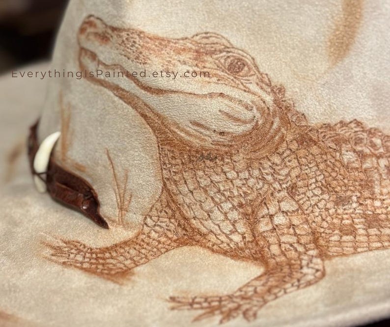 Pu&ograve; includere: Un cappello da cowboy beige con una dettagliata illustrazione marrone di un alligatore. Il disegno dell'alligatore &egrave; finemente disegnato sulla corona del cappello. Il cappello ha una fascia in pelle marrone e un accento di dente bianco. Il testo "EverythingIsPainted.etsy.com" &egrave; visibile.