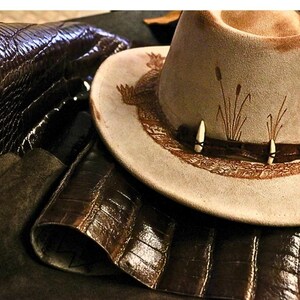 Pu&ograve; includere: Un cappello da cowboy in camoscio beige con una fascia decorativa con due accenti bianchi a forma di osso e una fascia in pelle marrone. Il cappello ha un design a tesa dettagliato ed &egrave; circondato da pelle marrone scuro.