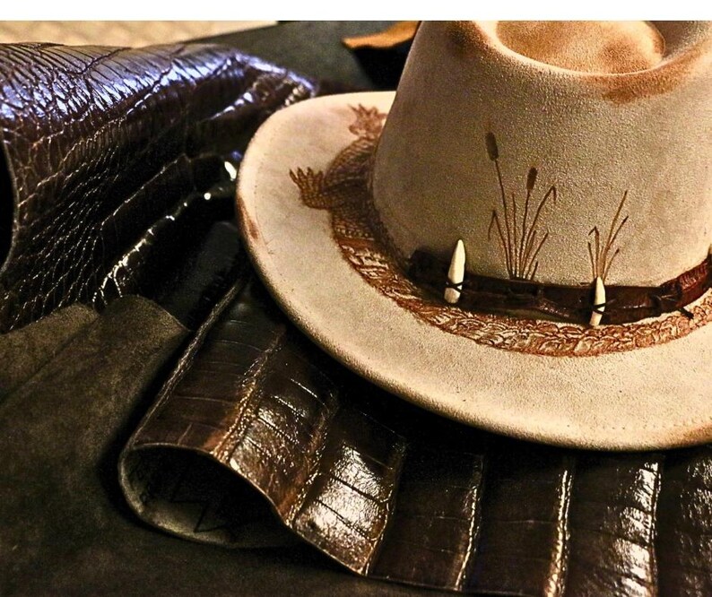 Pu&ograve; includere: Un cappello da cowboy in camoscio beige con una fascia decorativa con due accenti bianchi a forma di osso e una fascia in pelle marrone. Il cappello ha un design a tesa dettagliato ed &egrave; circondato da pelle marrone scuro.