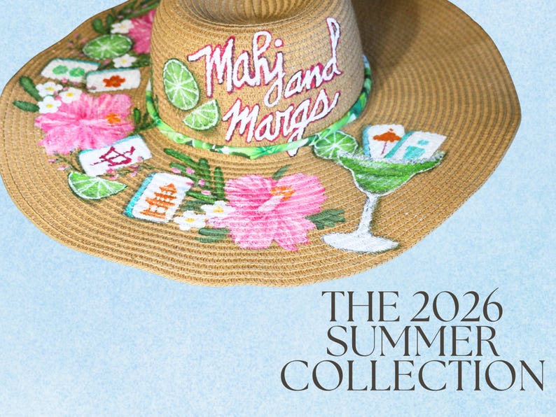 May include: A wide-brimmed straw hat with hand-painted tropical designs. The hat features pink hibiscus flowers, green lime slices, and the text "Mahi and Margs" on the brim. The hat also has a margarita glass design and the text "THE 2026 SUMMER COLLECTION".