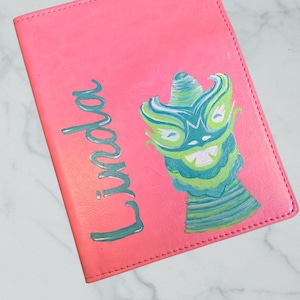 Puede incluir: Un diario de polipiel rosa con el nombre "Linda" en escritura turquesa. El diario presenta un diseño pintado a mano de una criatura turquesa y verde con una cara sonriente.