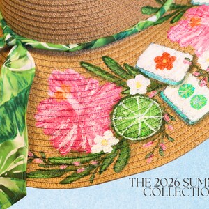 May include: A straw sun hat with a wide brim, adorned with pink hibiscus flowers, green leaves, and a lime slice design. A green and white patterned ribbon is tied around the hat. The text "THE 2026 SUMMER COLLECTION" is visible.