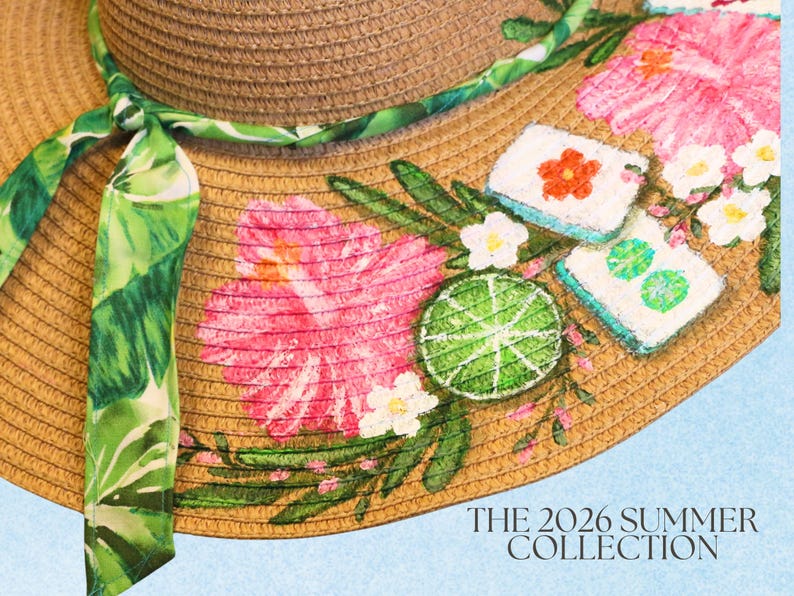 May include: A straw sun hat with a wide brim, adorned with pink hibiscus flowers, green leaves, and a lime slice design. A green and white patterned ribbon is tied around the hat. The text "THE 2026 SUMMER COLLECTION" is visible.