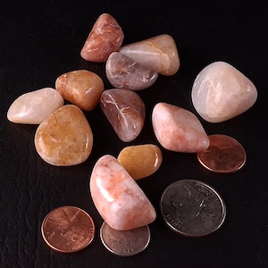 May include: A collection of polished, tumbled stones in shades of orange, peach, and cream, with a few translucent specimens. The stones are displayed with a penny and a quarter for size comparison. The background is black.
