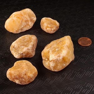 May include: Five smooth, rounded, light-orange stones of varying sizes are arranged on a textured black surface. A copper penny is next to one of the stones for size comparison. The stones have a natural, earthy appearance.