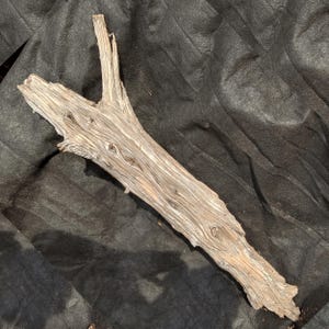 May include: Weathered driftwood branch with a Y-shaped split at one end. The wood is a light brown color with visible grain and knots, lying on a dark, textured surface. The branch appears to be naturally aged and worn.