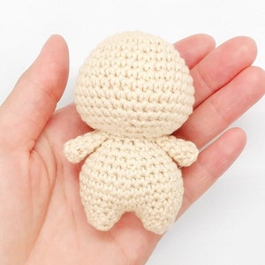 Puede incluir: Una muñeca de crochet con un cuerpo de color crema y sin rasgos faciales. La muñeca está sentada en la palma de una mano.